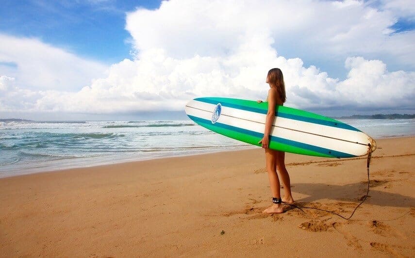 Woman holding a surf board facing the ocean on the beach