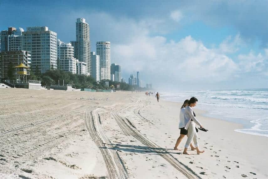Man and a woman walking down the beach towards the ocean on the right, with the Gold Coast skyscrapers in the background at the top of the beach