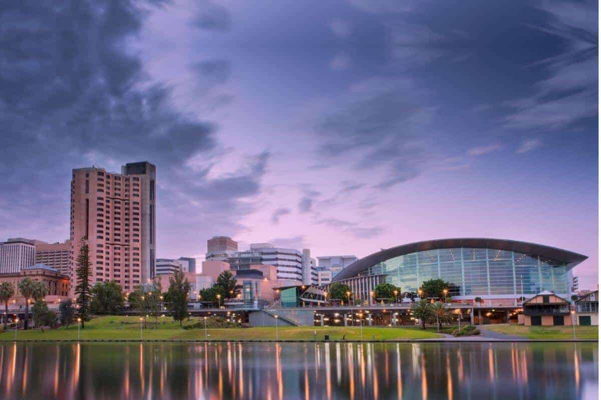 Adelaide CBD from the across the water of the River Torrens with a sky scraper on the left, buildings of the CBD in the middle and a large glass domed stadium building on the right. The sky is shades of purple, and the lights of the building are reflected in the river