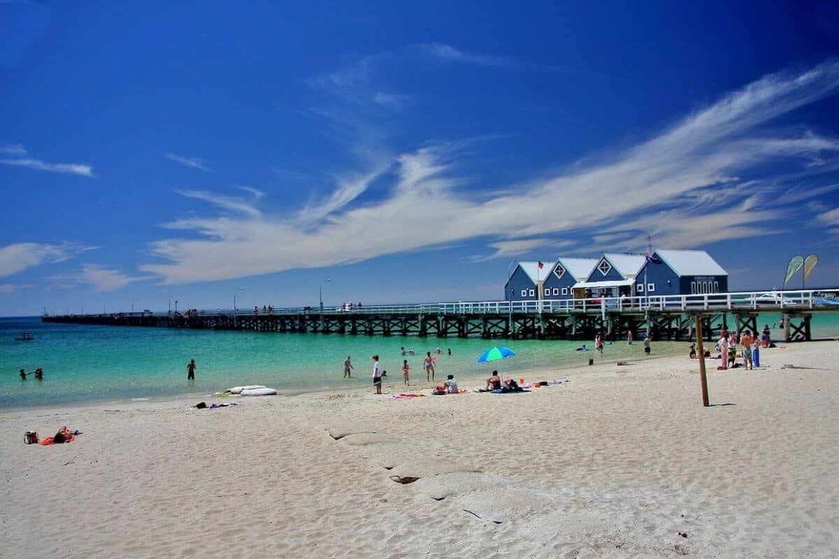 Top Travel Tips for Australia header image of Bussleton Beach with people sunbathing in front of the light blue ocean with the jetty in the background with 4 wooden beach huts on the jetty with no text overlay