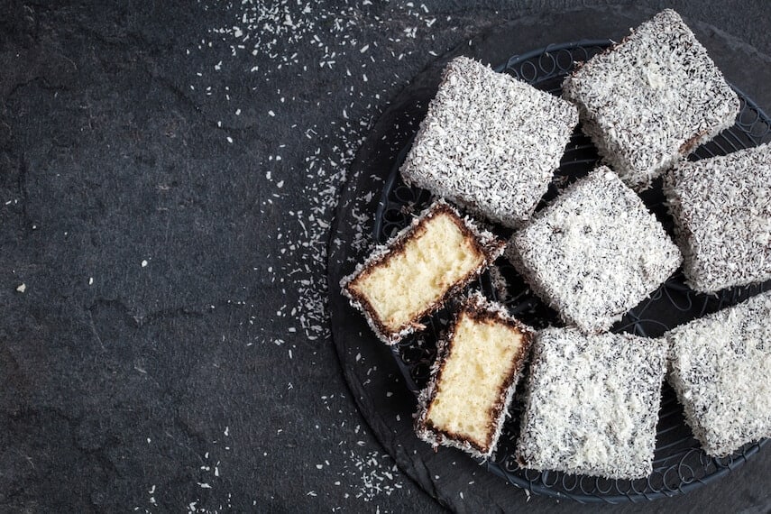 Dark brown squares of cake covered in desiccated coconut on a plate with one cut in half and facing up to expose the cake centre