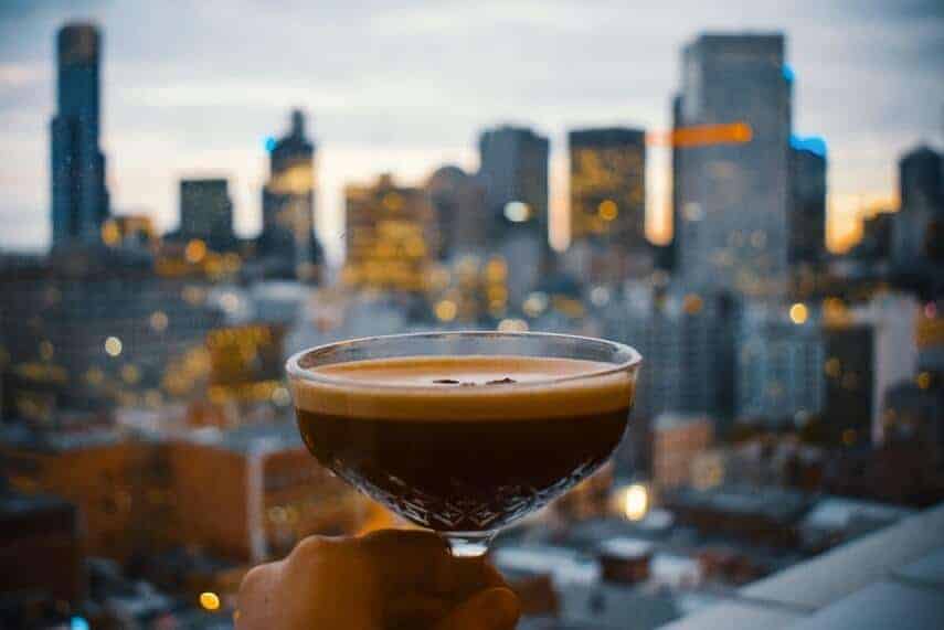 Woman holding an expresso martini in fouces in front of the Melbourne skyline at dusk