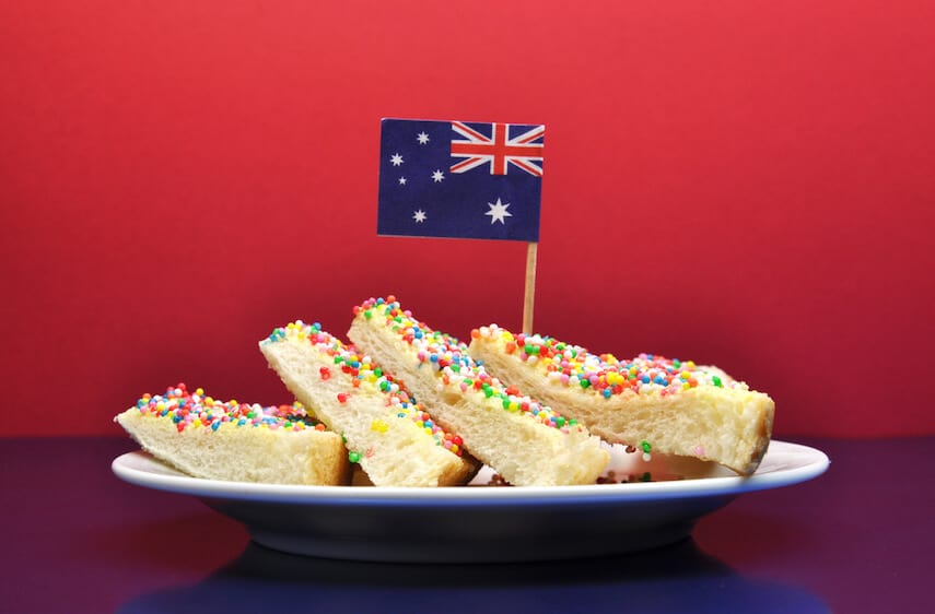 Slices of fairy break - white buttered bread covered in hundred sand thousands on a white plate, topped with a mini Australian flag in front of a red wall 
