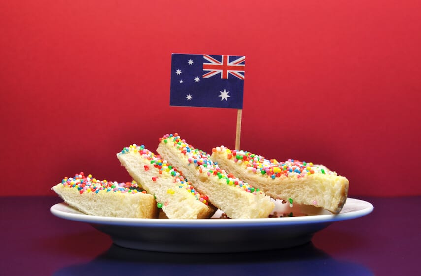 Slices of fairy break - white buttered bread covered in hundred sand thousands on a white plate, topped with a mini Australian flag in front of a red wall