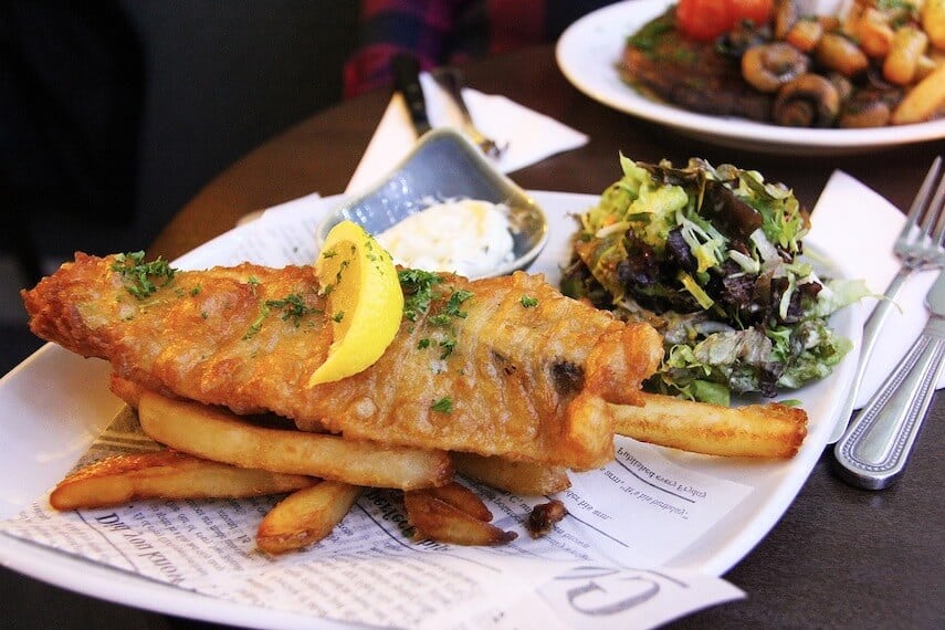 Beer battered fish on a bed of chips topped with a lime wedge and served with a side salad on a white plate