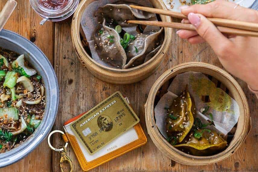 Gold amex card on a table surrounded by baskets of dumpings with a woman holding chopsticks above one