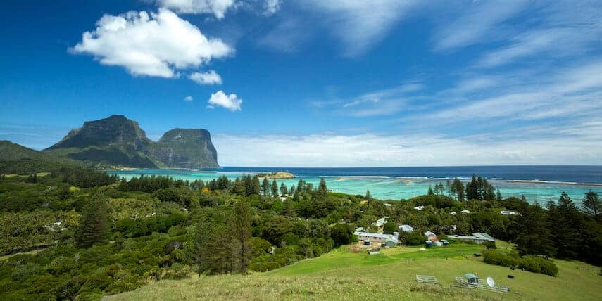 Panorama of Lord Howe Island with the green fields in the foreground, a few houses close to the coastline, the ocean in the background and the large mountains in the distance