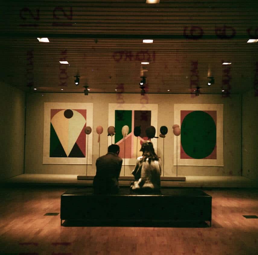 Inside the NGV with 3 artworks on the wall and two people sitting on a bench in the centre of the room looking at them