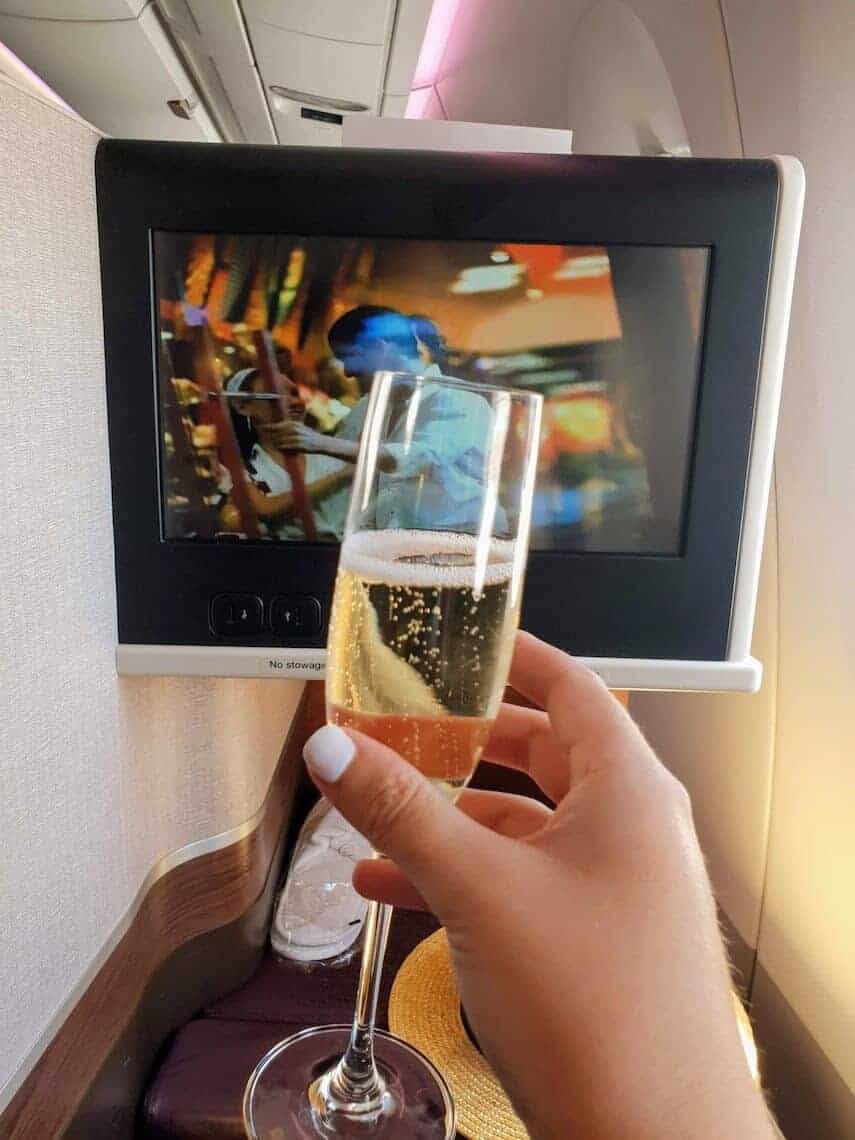 Woman holding a glass of champagne on a 30 degree angle in front of a large TV screen in the back of a business class seat on an airplane