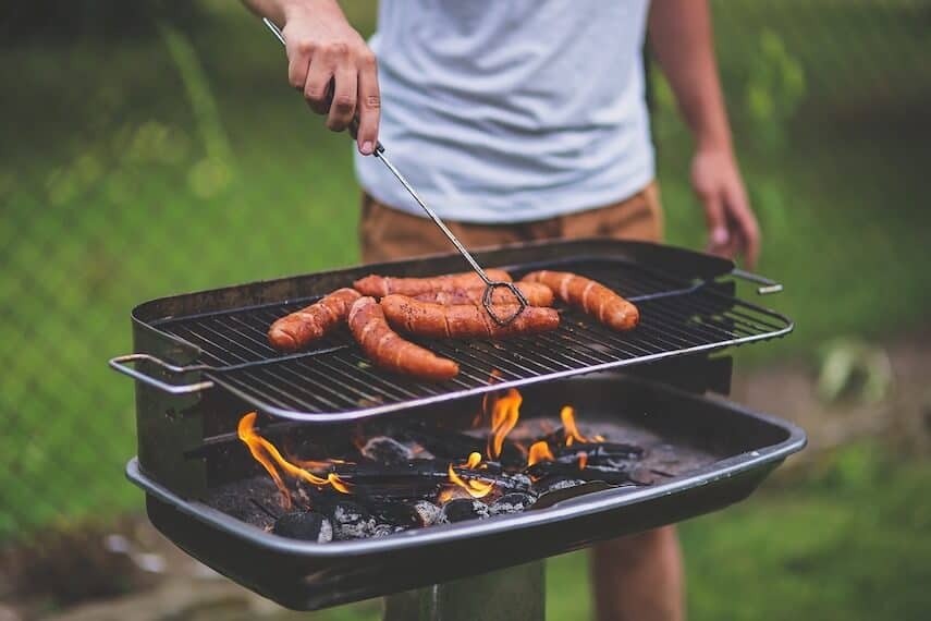Sausages on a coal fired BBQ, a man standing behind with a pair of tongs to turn the sausages