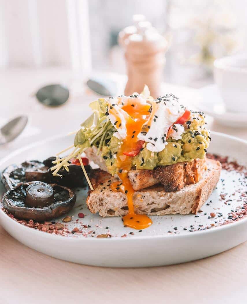 Elaborate smashed avocado on sourdough toast with a poached egg broken on top on a plate with fried mushrooms