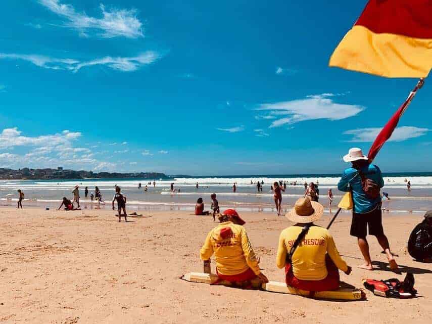 Busy Australian beach with two surf rescue lifeguards sitting on the sand next to the yellow and red safe swim zone flags