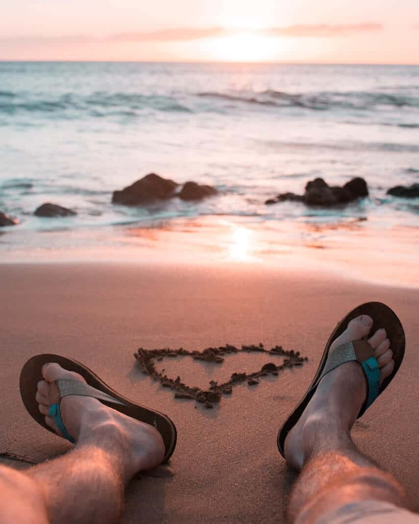 Mans legs wearing flip flops on the sand pointing towards the ocean with the sun setting in the background