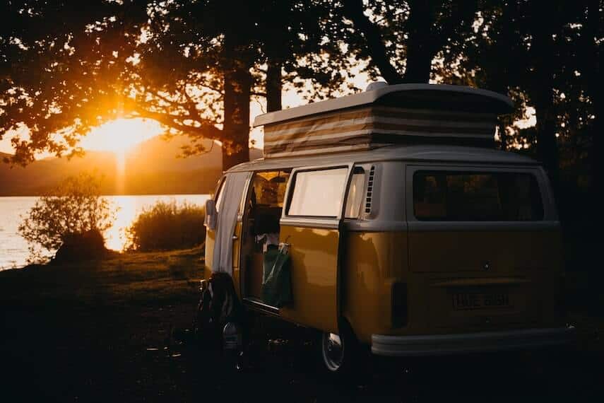 VW Combi Van with pop top parked next to a lake, with trees behind it at sunset