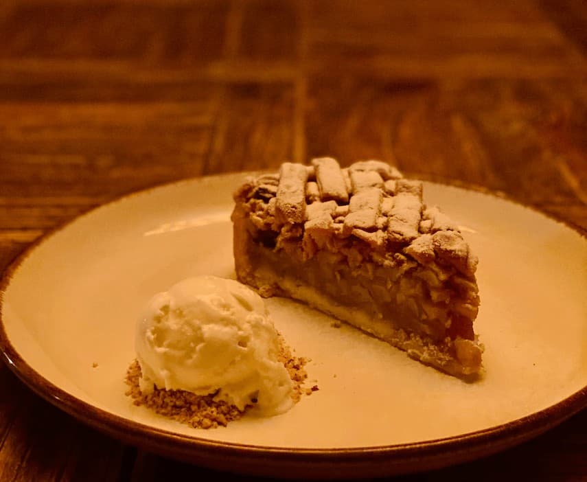 Slic of Apple Pie on a white plate with a ball of icecream on a bed of crumbs next to it. The plate sits on a wooden table
