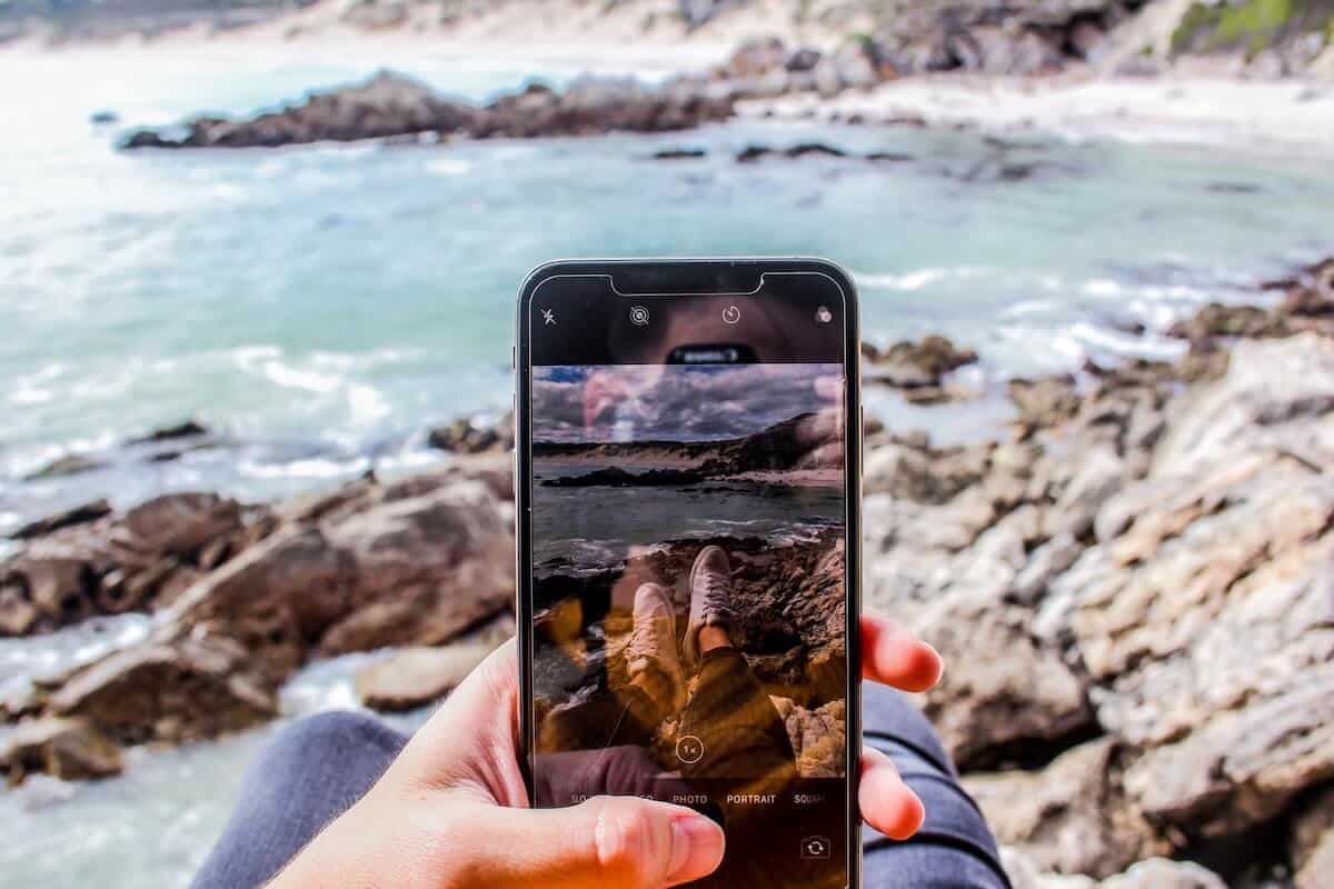 Australia Travel Apps Header image of a man sat on rocks next to the water looking at his phone