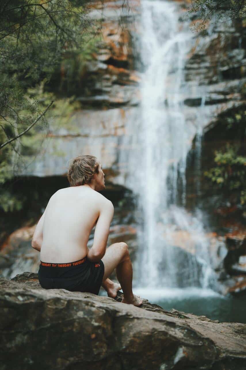 Man sat wearing board shorts and no t-shirt on a rock in front of Minnehaha Falls in the Blue Mountains