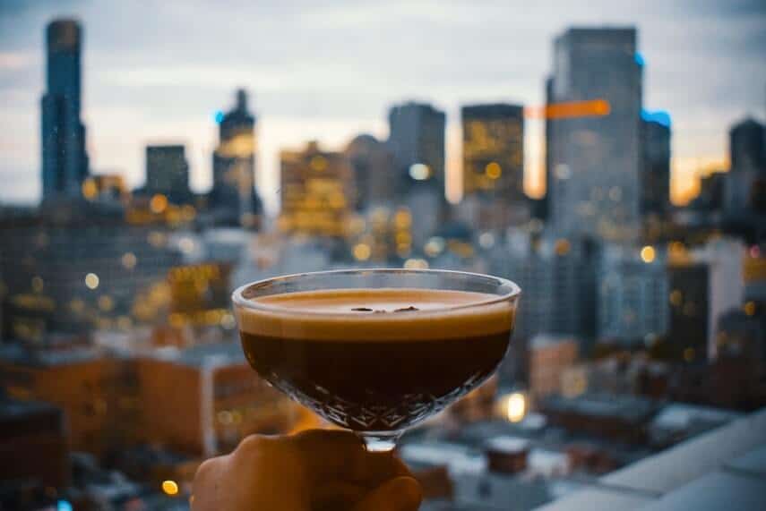 Woman holding an expresso martini up in front of the Melbourne skyline from a rooftop