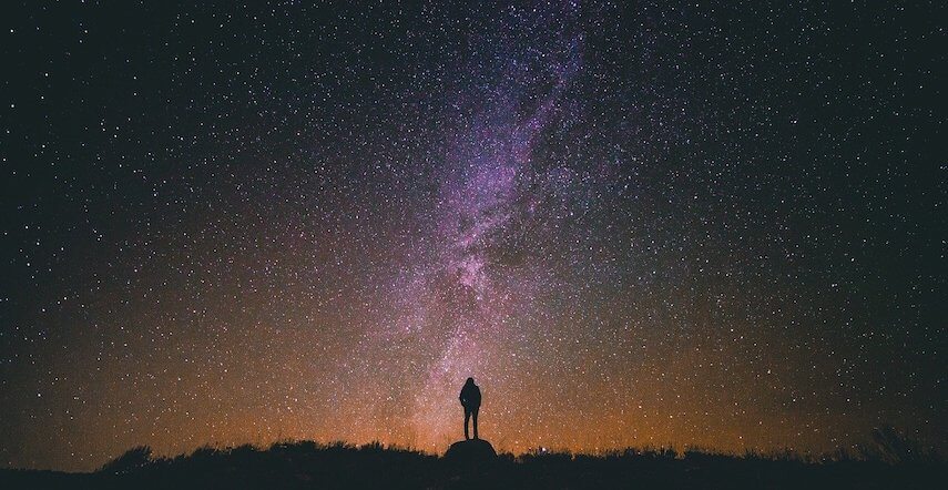 Man standing in silhouette in front of the milky way at sunrise, the sky nearest the horizon blends into the purple of night at the top