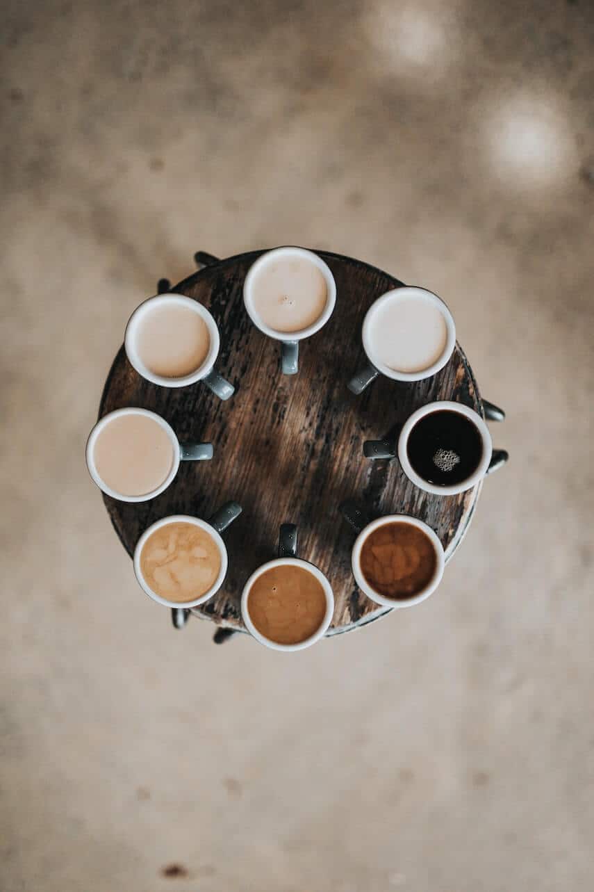 Top down shot of a round dark wood table with 8 mugs of coffee each a different colour lined up around the edge of the table