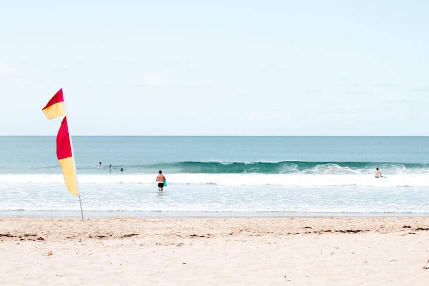 Light beige sand with waves crashing onto the shore, and the red and yellow lifeguard flag planted into the sand on the left of the image