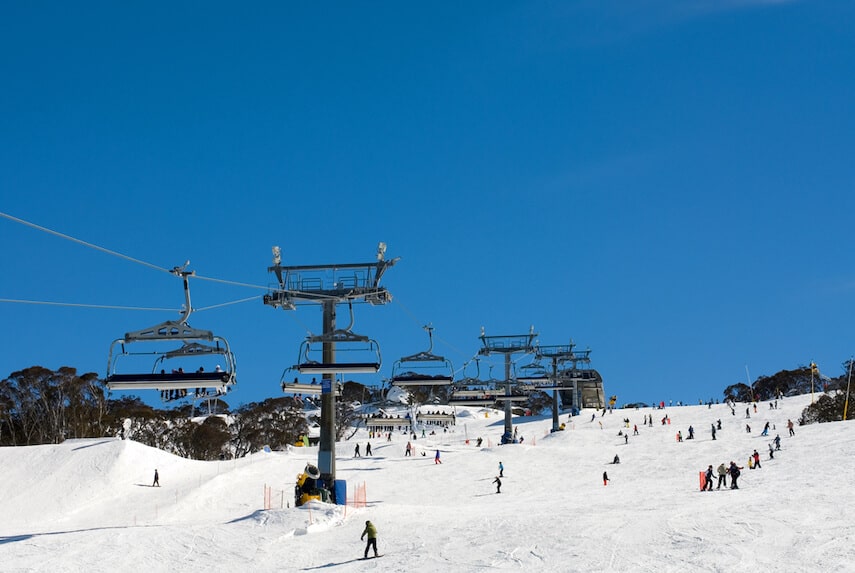 Perisher Ski Resort NSW view of a ski slope, lift station building at the top and the 8 person chairlift running up the middle. People are skiing and snowboarding down the slope