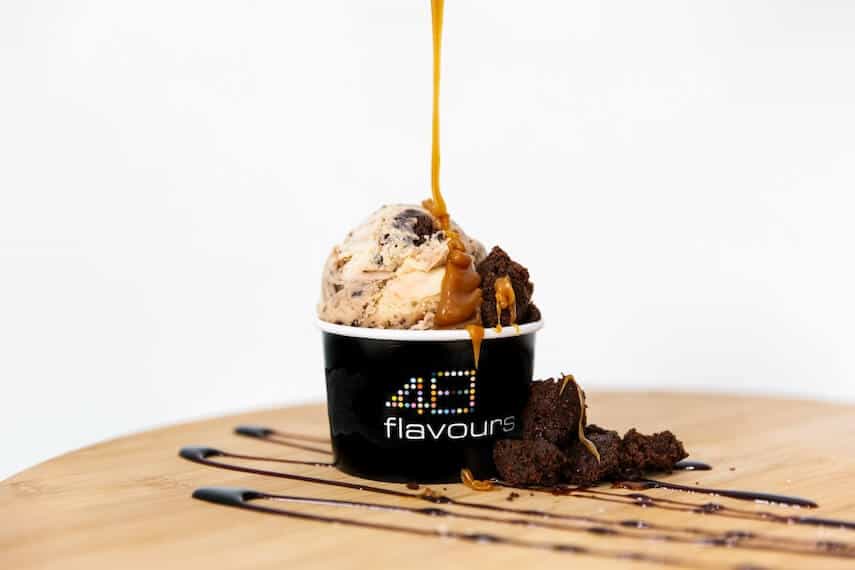 48 Flavours branded ice cream tub on a wooden board with caramel flavoured ice cream being drizzled with a dark caramel sauce