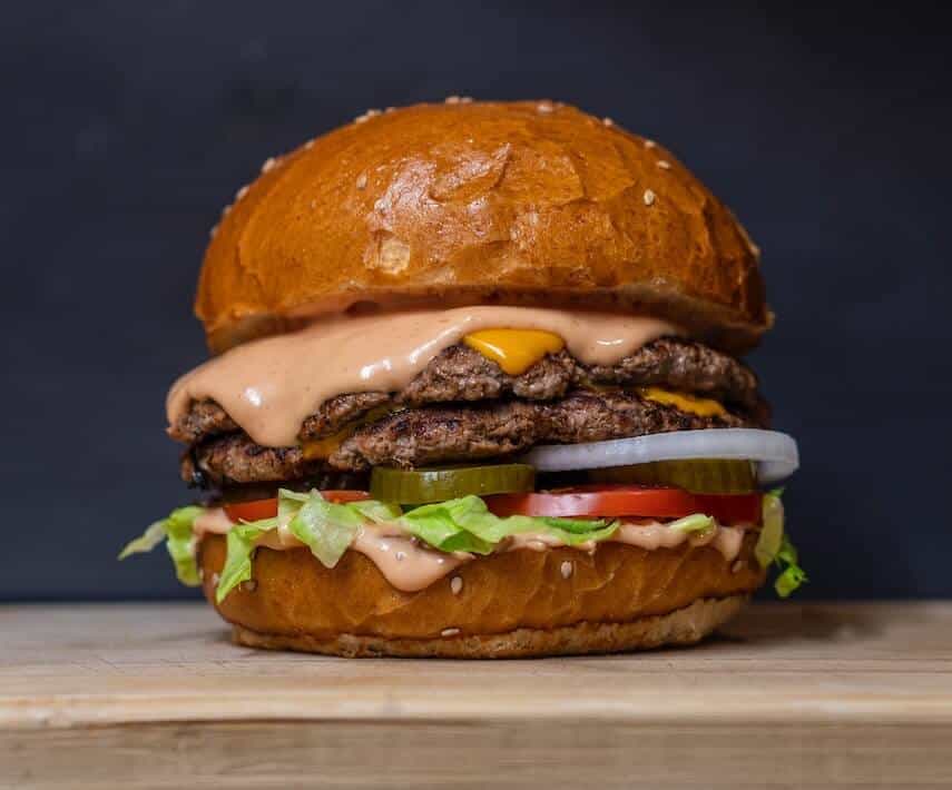 side view of a double beef patty burger in a seeded bun with burger sauce and cheese dropping over the top patty and assorted salad items underneath