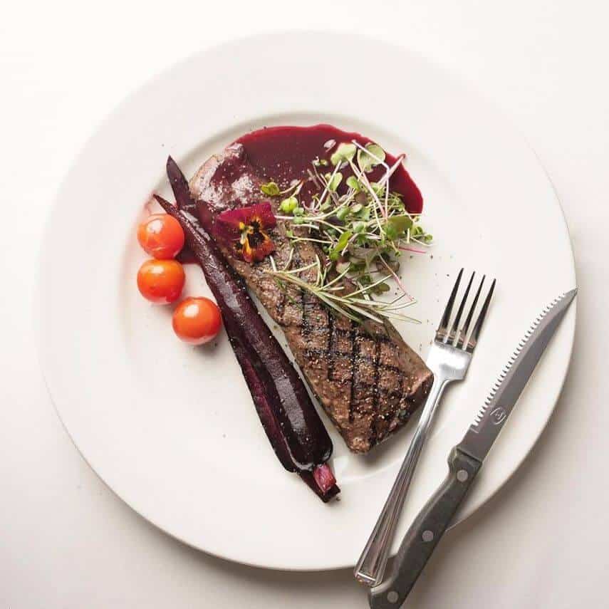 Venison with root vegetables on a white plate, a knife and fork resting on the side of the plate