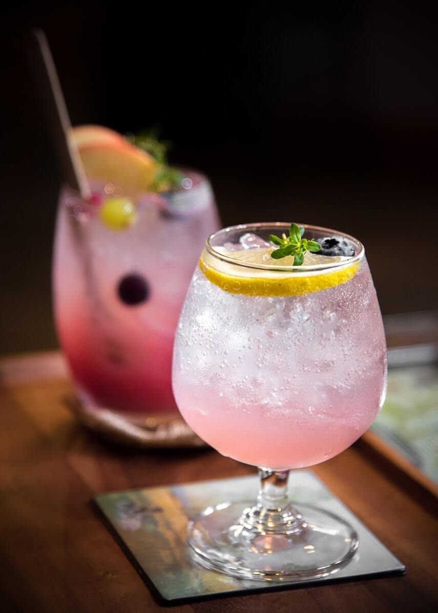 Two pink coloured cocktails on a wooden board, the one at the front is topped with a lemon wedge, the one at the back garnished with a blackberry and orange wedge with a metal straw