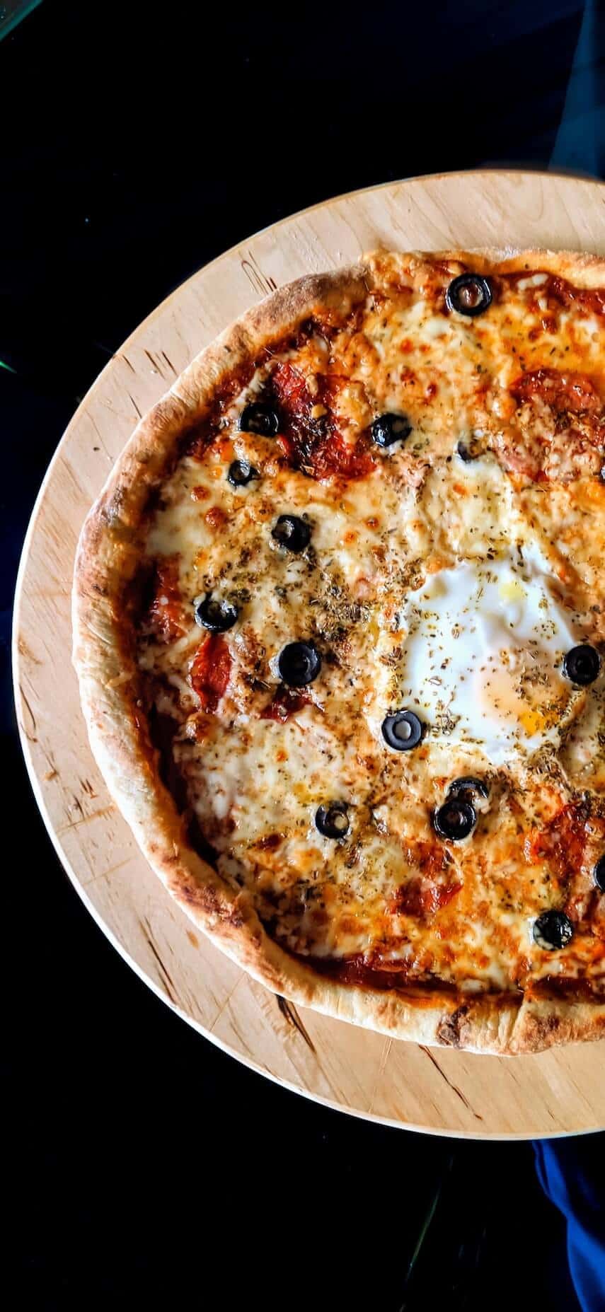 Top down shot of a pizza with cheese and olives on a circular wooden board on a black table