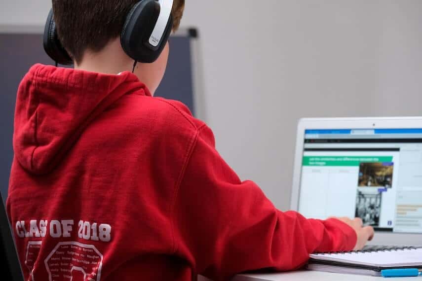 Young boy wearing a red hoodie and black headphones sitting with his back to the camera facing a laptop, remote learning.