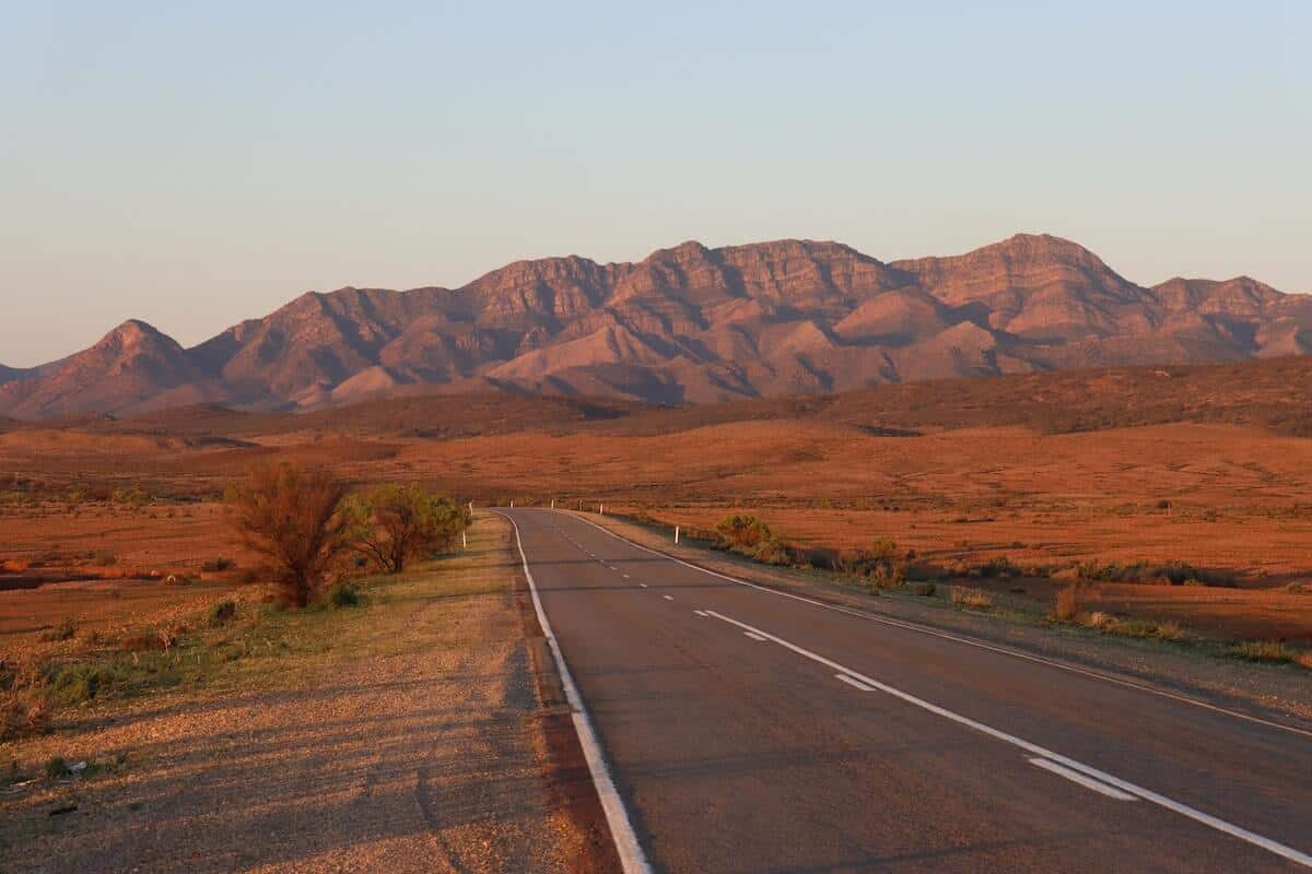 Bucket-List Worthy Road Trips in Australia cover photo of a road surrounded by red earth leading towards a large red rock formation in the distance