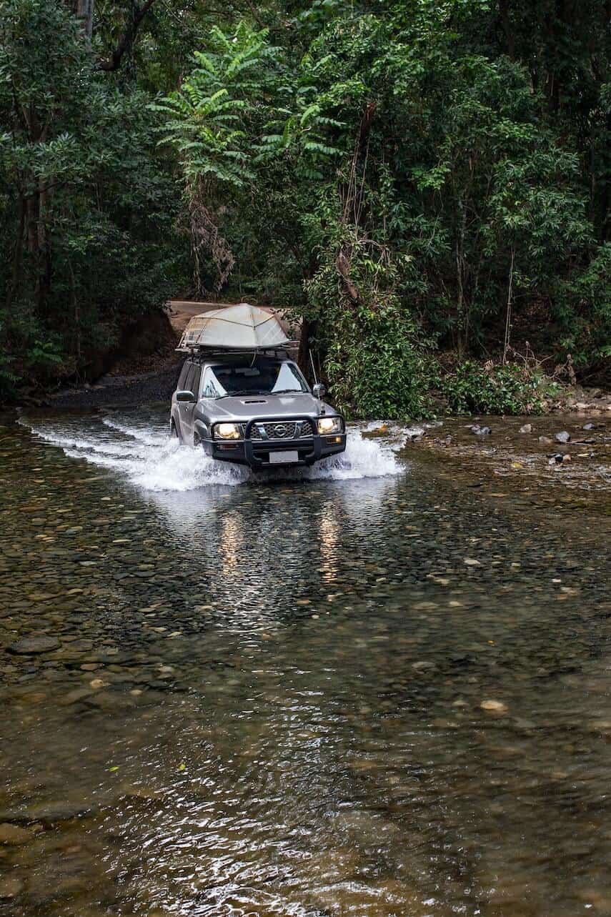 4x4 Car with an upside down tin boat on the roof driving through a shallow river surrounded by rainforest