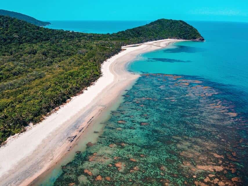 Green Rainforest borders a White sand beach which meets a shallow reef surrounded by clear blue ocean